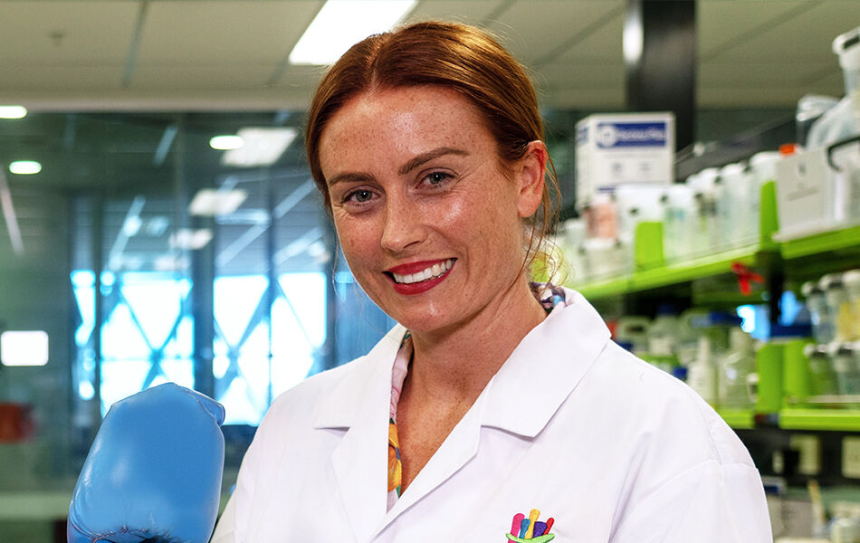 Professor Hannah Wardill smiling in labcoat with blue boxing gloves