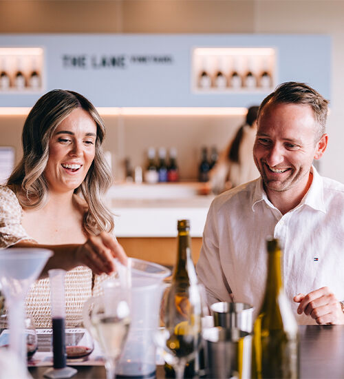 Woman and man smile as they pick up mixing tools on table with wine bottles